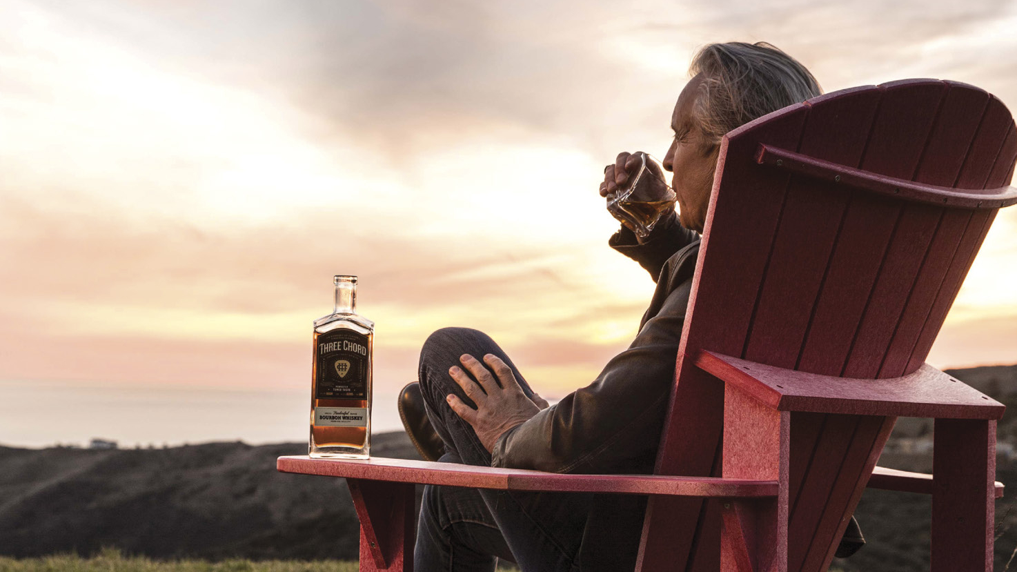 Neil Giraldo sipping whiskey in a red Adirondack chair at sunset beside a bottle of Three Chord Bourbon