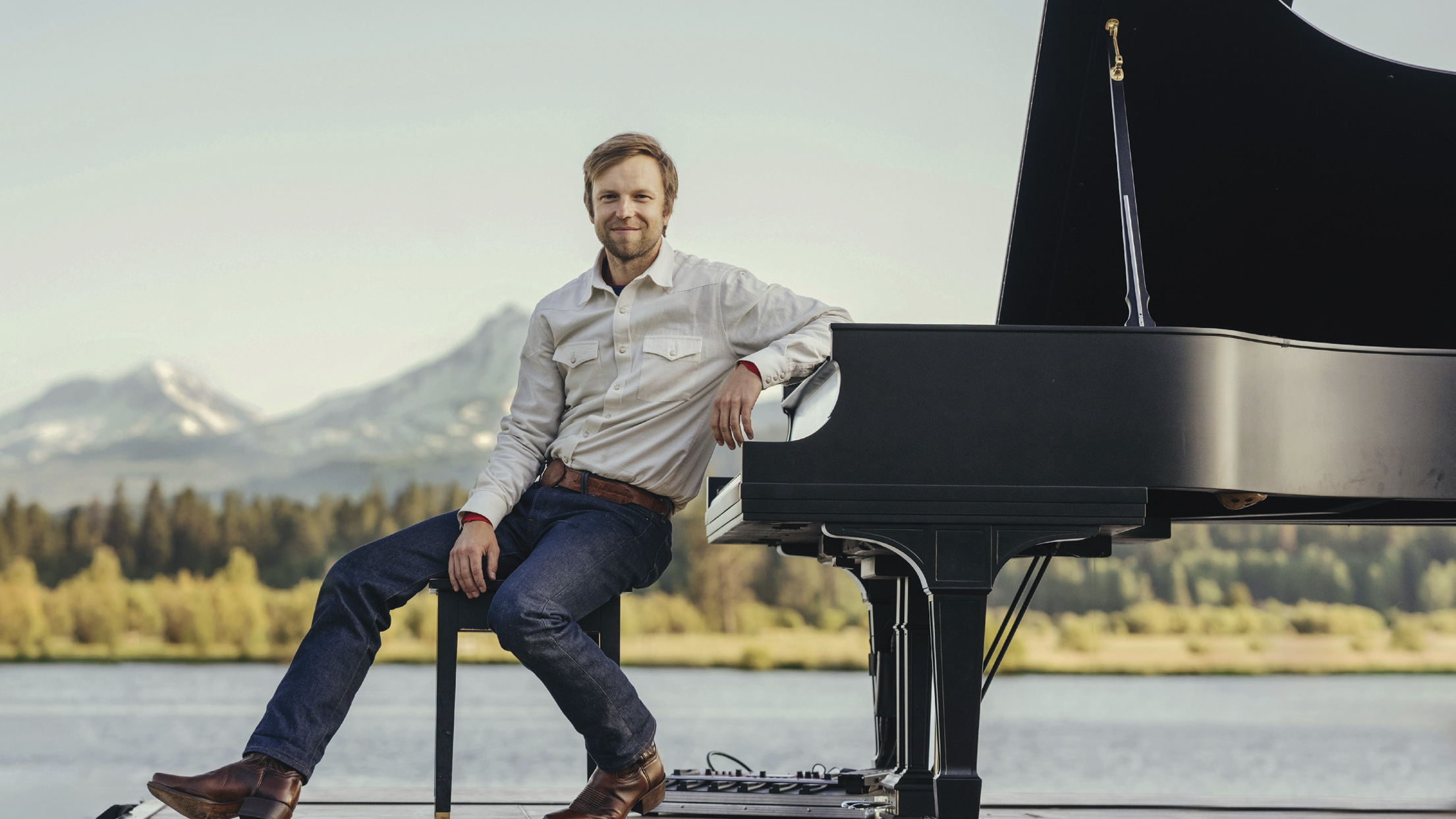 Pianist Hunter Noack seated beside a grand piano on a lakeshore with snowcapped mountains behind him