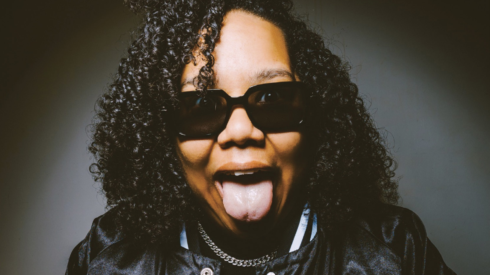 Rapper Mama Duke in sunglasses and a black jacket sticking out her tongue playfully