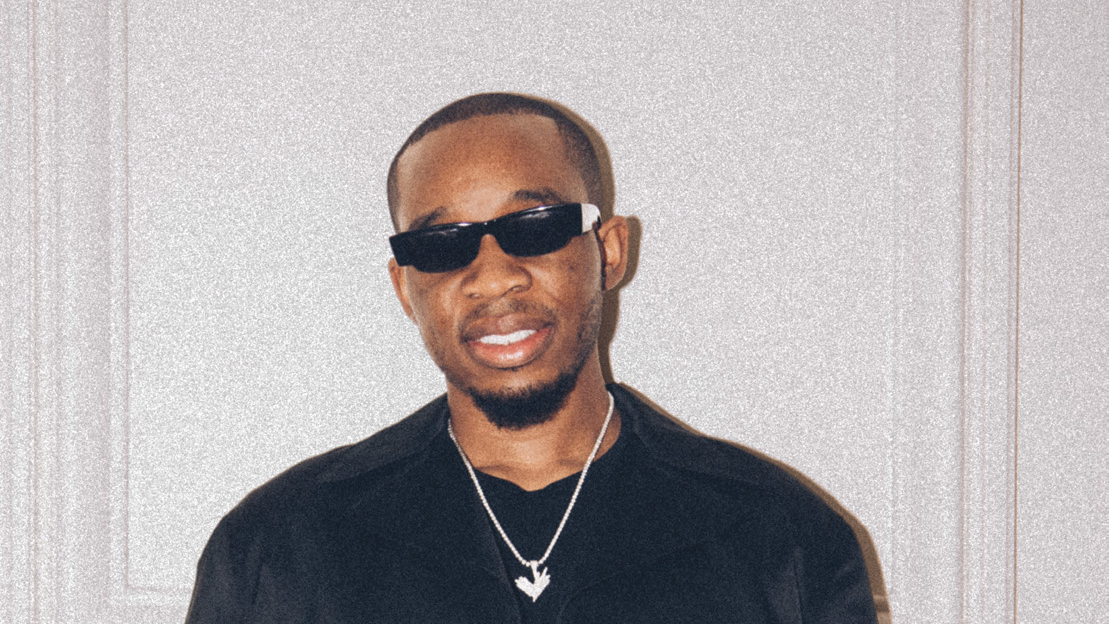 Tunde Balogun in sunglasses, a black jacket and a chain necklace against a white paneled wall
